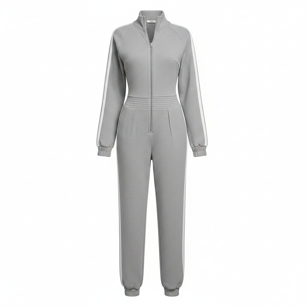 A woman models a light gray zip-up tracksuit with white athletic stripes down the sleeves and legs, styled with white sneakers and a brown patterned tote bag against a textured gray concrete wall.