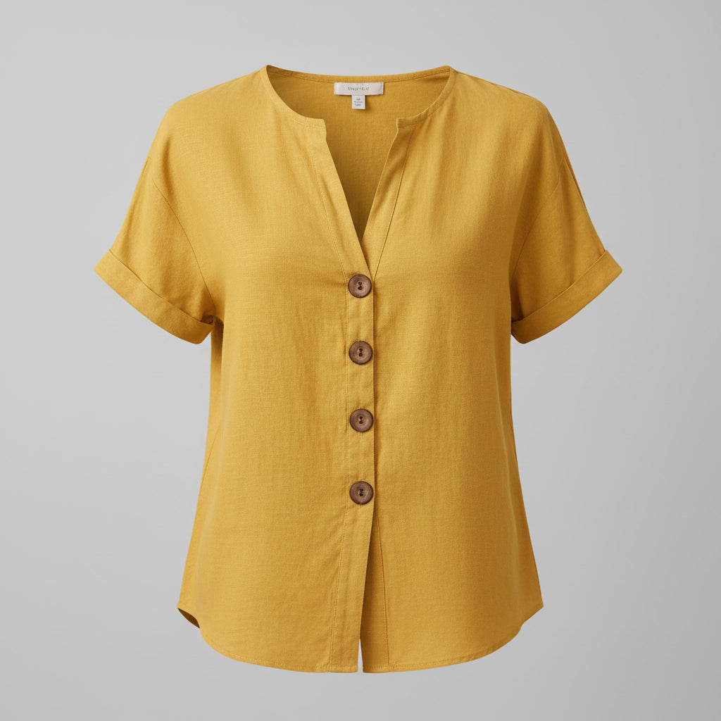 A woman wearing a mustard yellow linen short-sleeve button-up top and cream pants, accessorized with bracelets and a necklace, standing in a styled indoor setting with woven decor.