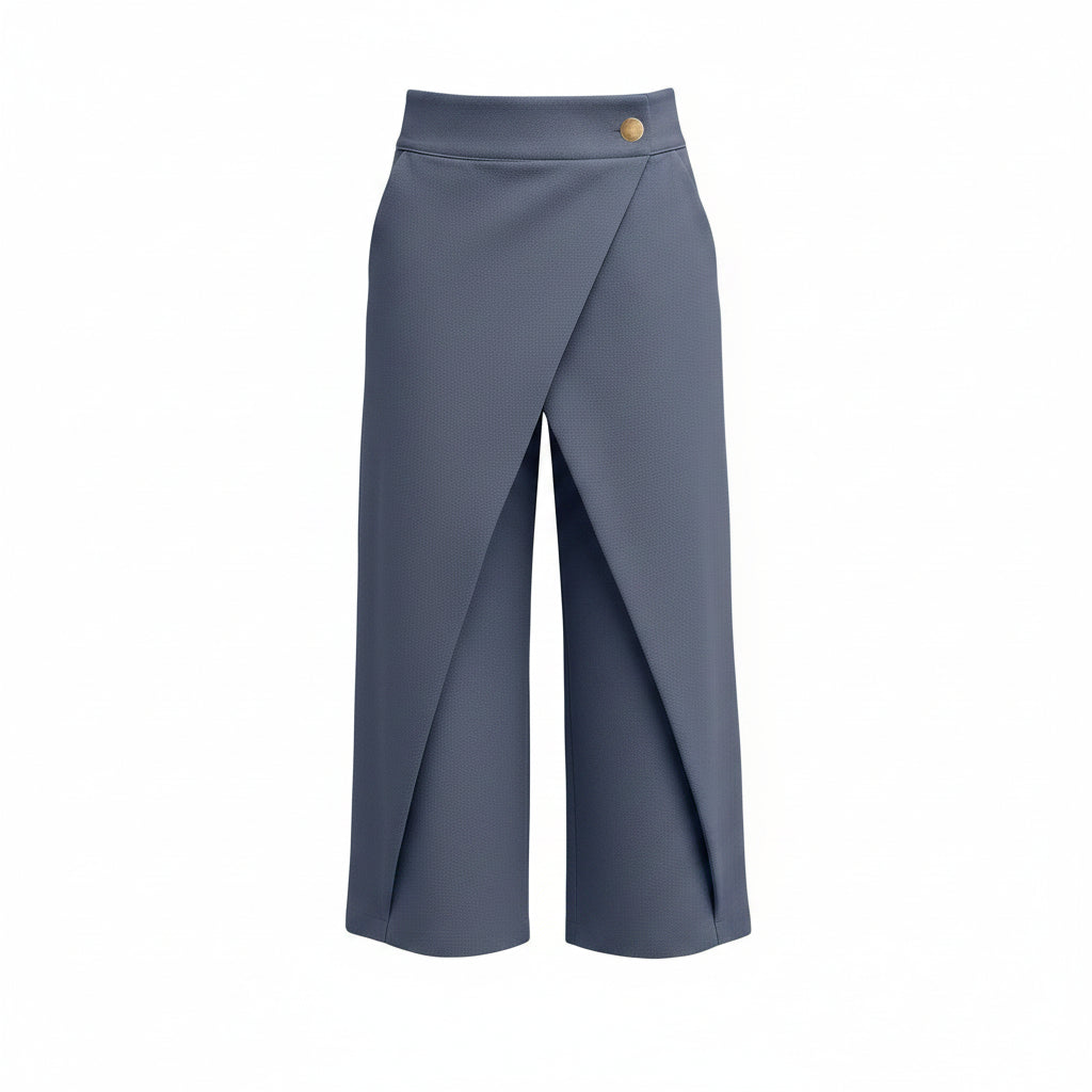 A model wearing slate blue wrap-style cropped trousers with a single gold button at the waist, paired with a white long-sleeve blouse and silver heels, against a solid gray studio background.