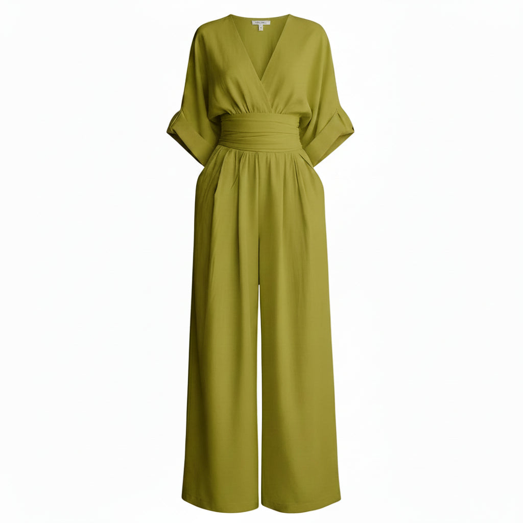 Woman wearing a green jumpsuit standing next to a textured wall and decorative vase.
