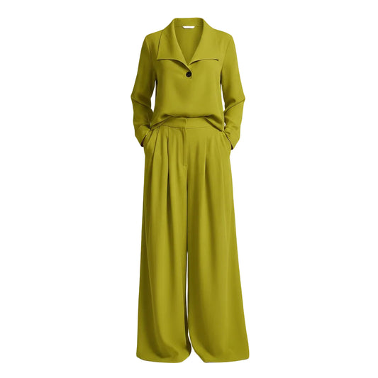 A woman wearing a bright chartreuse green two-piece set consisting of a long-sleeve cowl-neck top and matching wide-leg pants, standing on a sandy beach with a thatched-roof structure in the background.