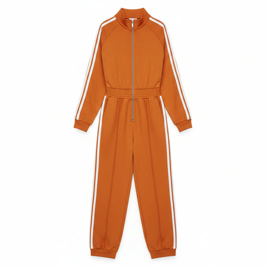 A woman models a matching orange zip-up sweatshirt and wide-leg sweatpants set with white side stripes, styled with white sneakers and a brown patterned tote bag against a gray concrete wall.