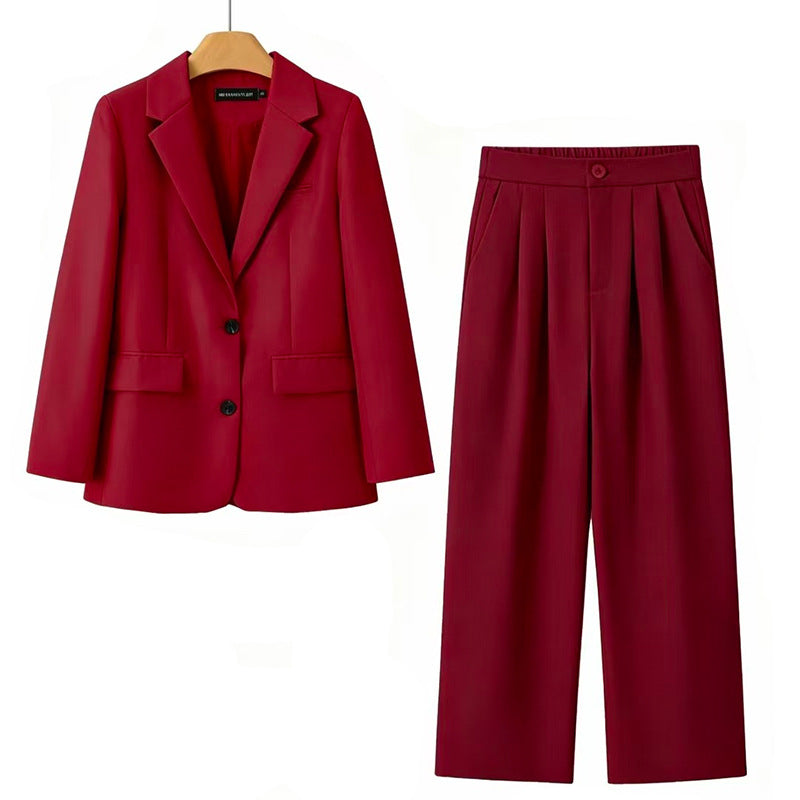 A two-piece women's suit set in a rich burgundy red, featuring a single-breasted blazer on a wooden hanger and matching wide-leg pleated trousers, displayed against a solid white background.