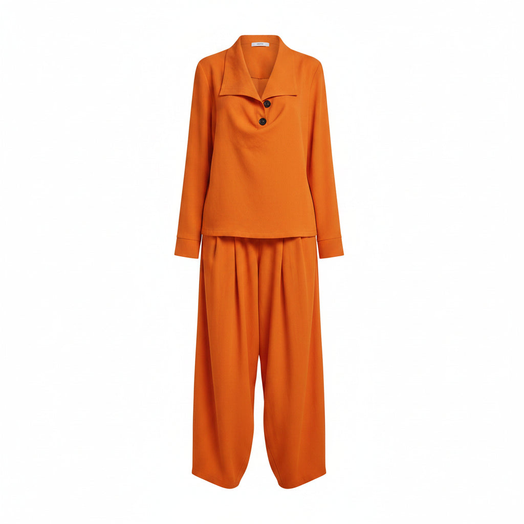 A person wearing a bright orange two-piece outfit with a long-sleeve collared top and matching loose wide-leg pants, standing on sand in front of a white wall and thatched roof.