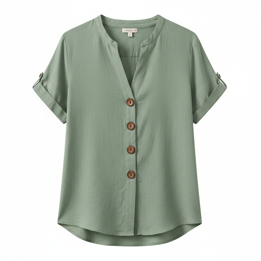 A woman wearing a sage green short-sleeve linen button-up blouse with brown buttons, paired with cream pants, shown in a styled indoor setting.