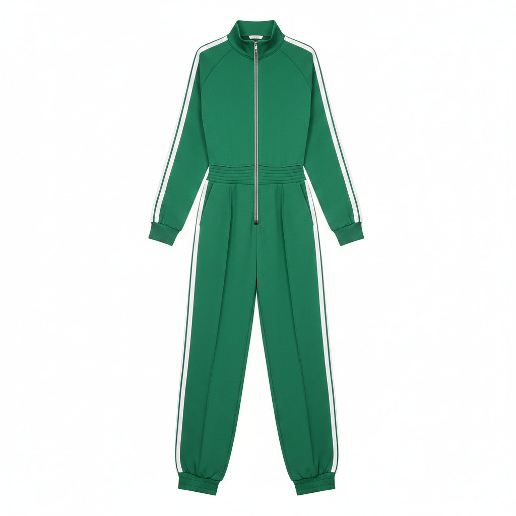 A woman models a bright green two-piece tracksuit with white side stripes, featuring a half-zip long-sleeve top and matching wide-leg pants. She is holding a brown monogram tote bag and wearing white sneakers, posed against a textured gray concrete wall.