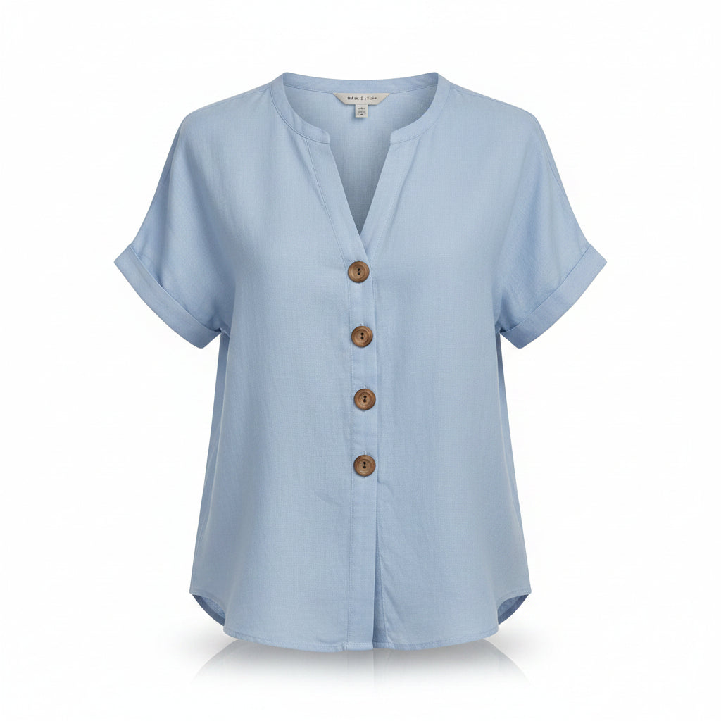 A woman wearing a light blue short-sleeve linen button-up blouse with brown buttons and cream pants, styled with jewelry in a warm, bohemian indoor setting.