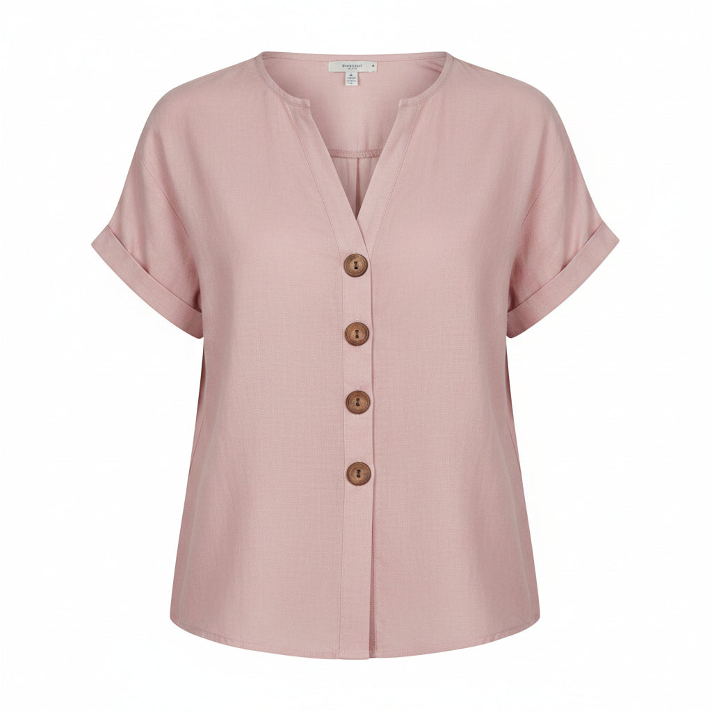 A woman wearing a light pink short-sleeve linen button-up blouse with brown buttons, paired with off-white pants, in a warm, bohemian-style room setting.